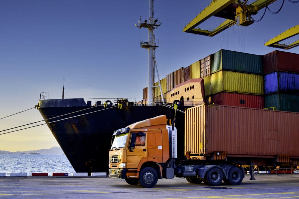 Truck carrying forty foot container leaving port terminal with ship and quay crane on the background. Seaport operation activities, container shipping, and logistics.